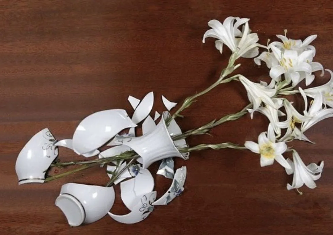 Un vase de porcelaine blanche brisé, des lys répandus sur une table en bois